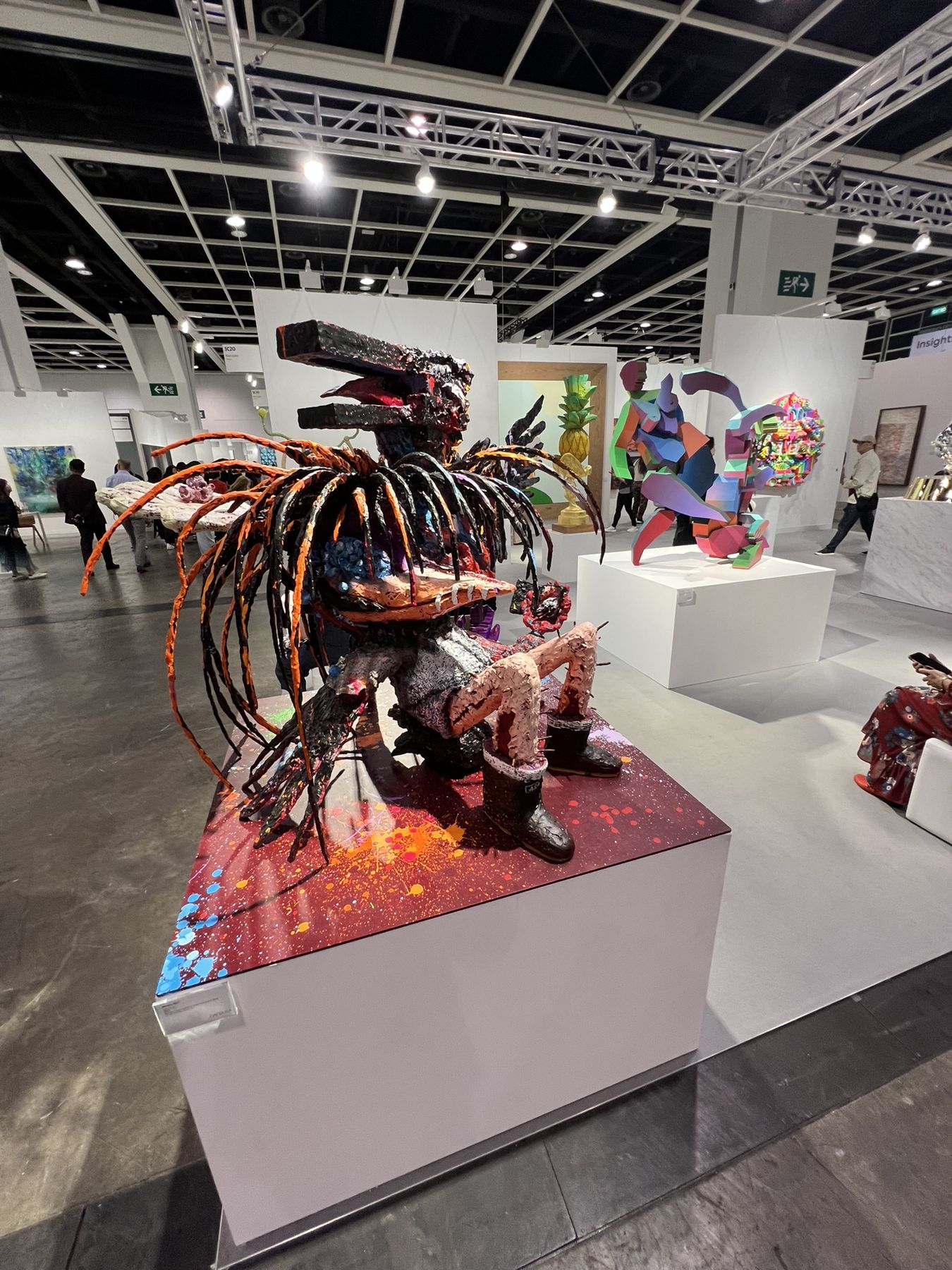 A busy art fair hall with a large mixed-media sculpture on a plinth in the foreground